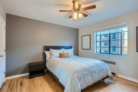 Bedroom with wood floor, plush bed with headboard, bedside drawers, ceiling fan, and large window.