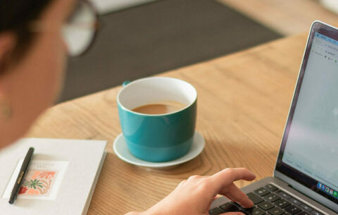 Person working on laptop at desk with coffee cup and notebook.