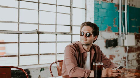 Person with sunglasses and cell phone sitting at wood table with iced coffee.
