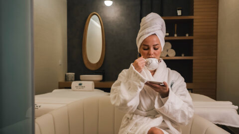 Person in robes and head towel relaxing with cappuccino at spa.